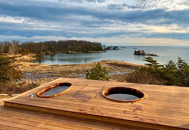 Wooden deck with two filled hot tubs overlooking a coastal landscape.